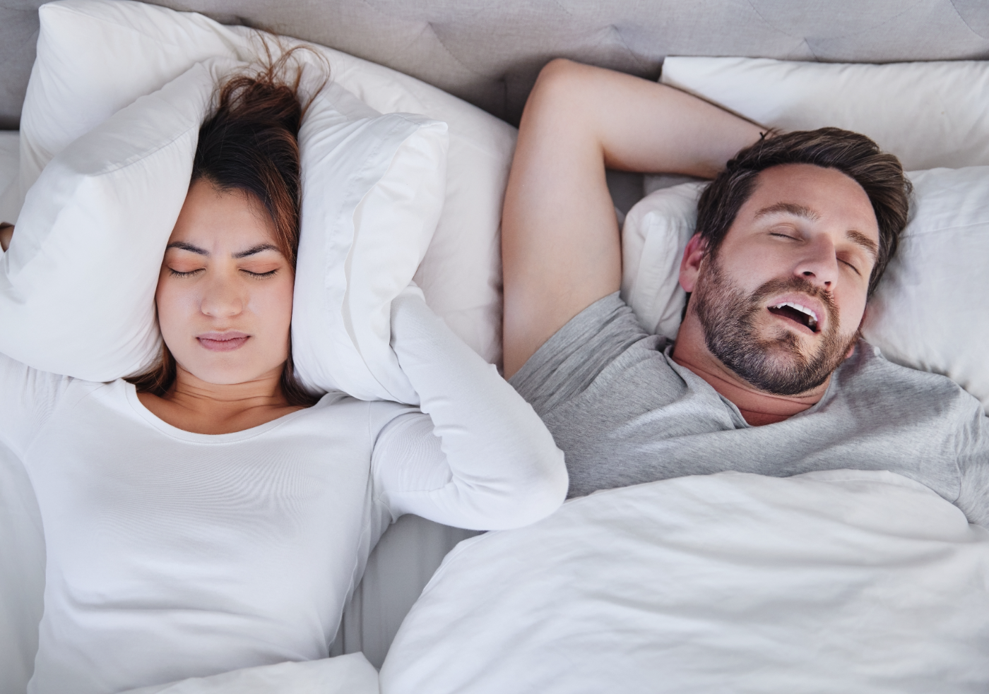 A man snoring with his mouth open while his partner covers her ears with a pillow