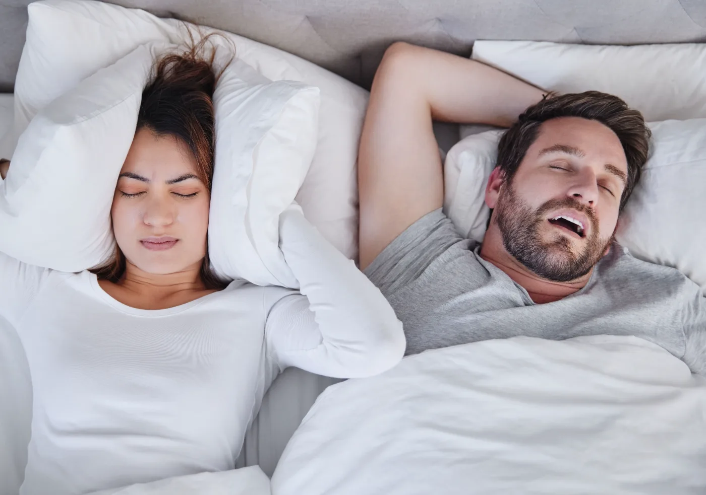 A man snoring with his mouth open while his partner covers her ears with a pillow